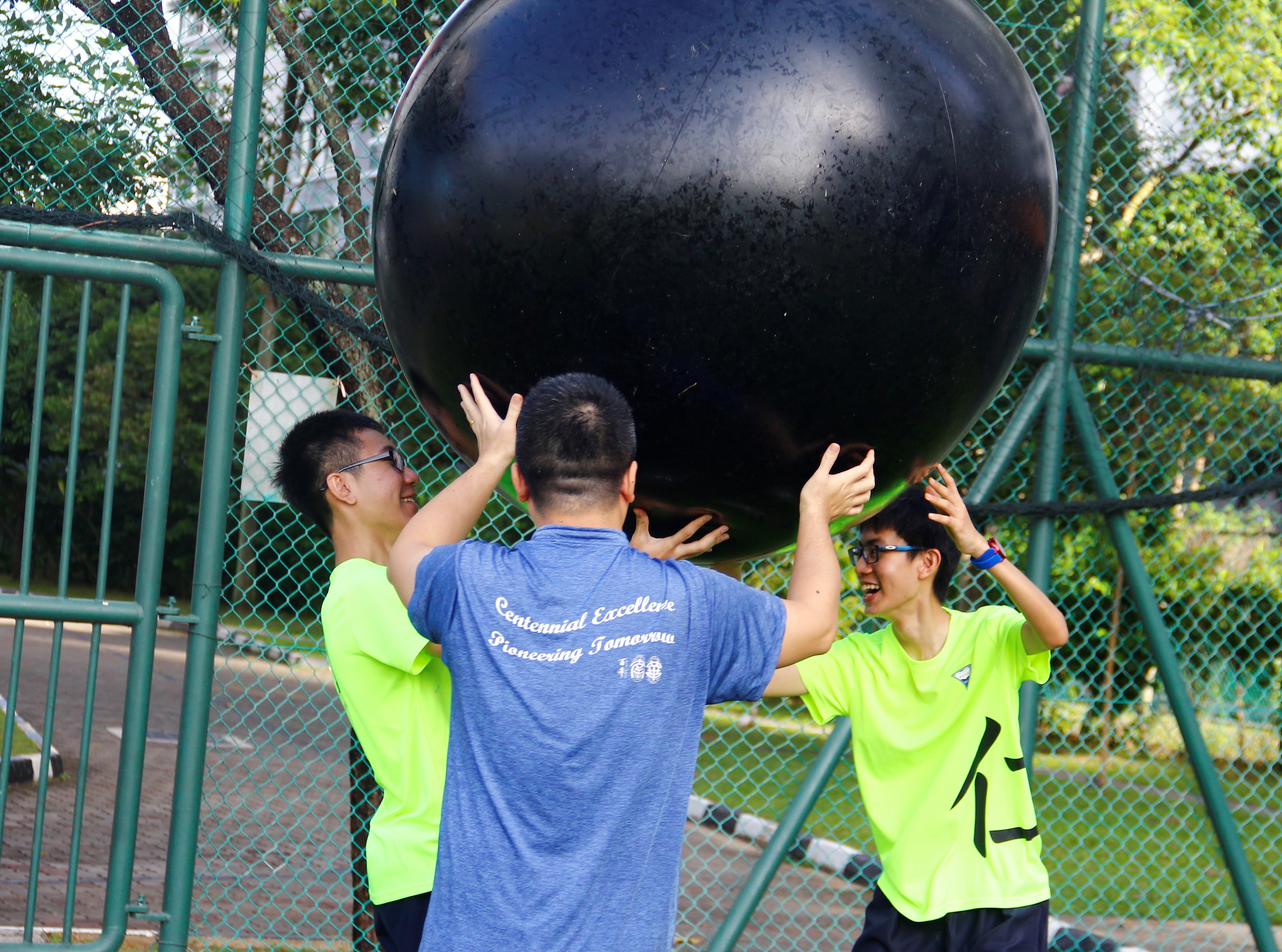 Three young people in athletic shirts lift a large black ball together outdoors near a green chain-link fence.
